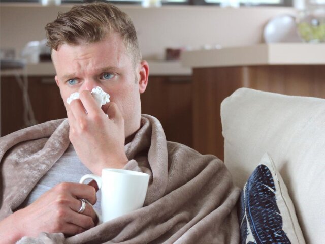 man-with-runny-nose-drinking-hot-drink-at-home-under-blanket-on-sofa