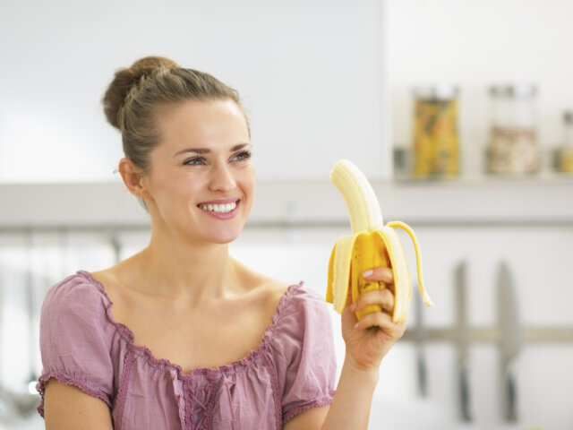Happy young woman eating banana in kitchen
