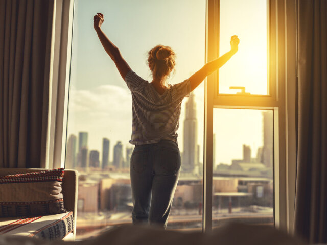 happy woman stretches and opens curtains at window in morning