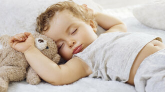 Sleeping baby boy arms raised up with a Teddy bear on a white blanket