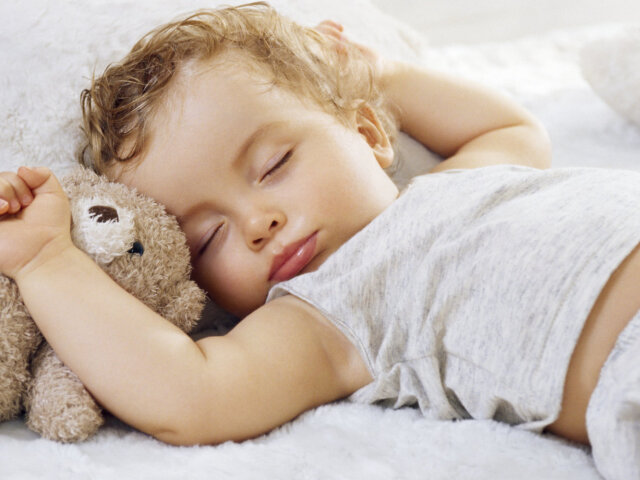 Sleeping baby boy arms raised up with a Teddy bear on a white blanket